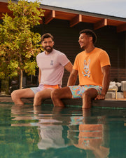 Two men sit on the edge of a pool with their feet in the water. Both are wearing T-shirts and Psycho Bunny MENS WINTON PRINTED SWIM TRUNK - B6W915A2PO featuring HD-printed bunny designs. One man wears a lavender shirt, the other an orange shirt. They smile and converse, with a modern building and tree in the background.