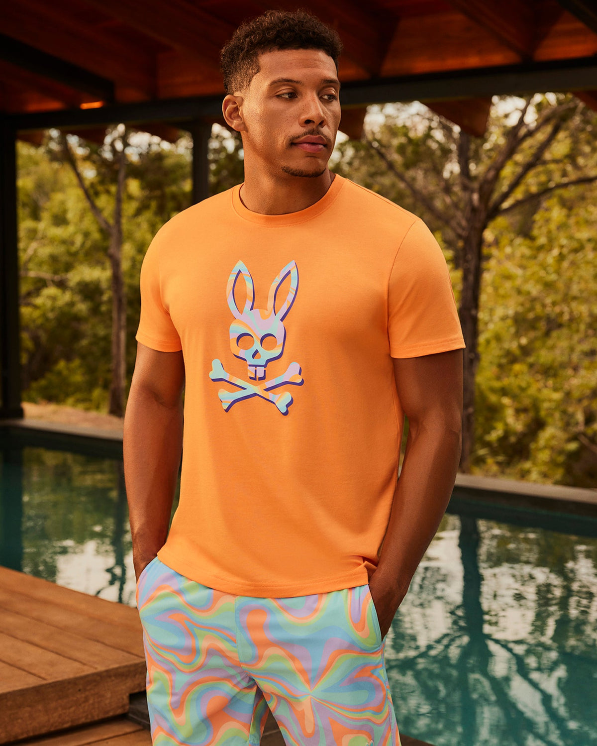 A man wearing an orange t-shirt with a blue silicone bunny patch stands poolside, wearing Psycho Bunny swim trunks featuring colorful patterns (MENS BLOOMINGTON SWIM TRUNK - B6W941A2PO), with a wooden structure and trees in the background.