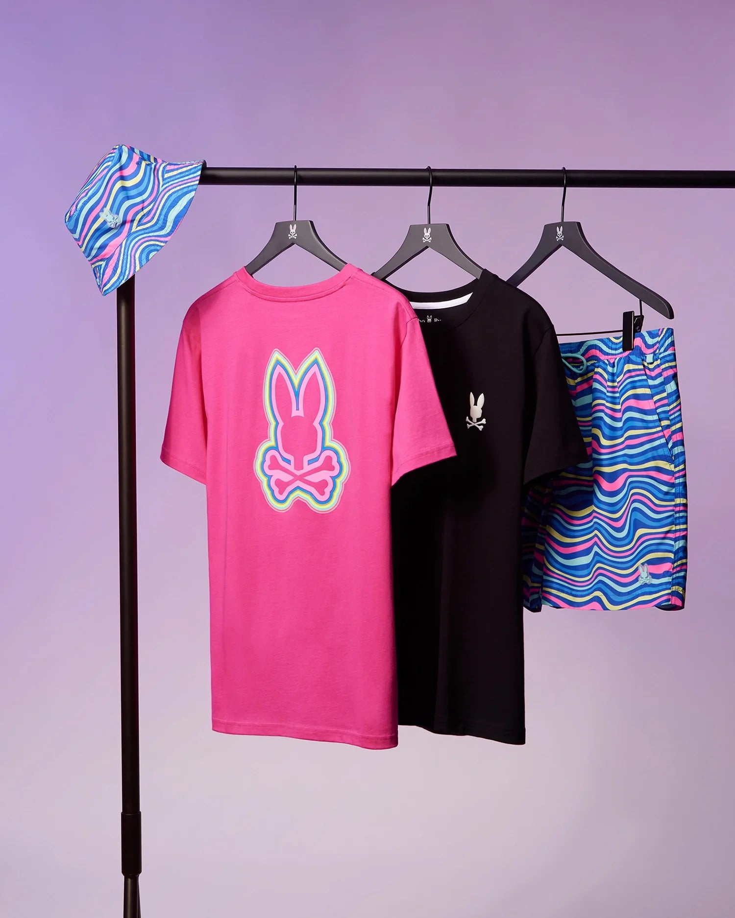 A clothing rack features a colorful bucket hat with a wave-like pattern, a pink t-shirt with a bunny skull graphic on the back, a black t-shirt with a small bunny skull on the front, and the Psycho Bunny MENS CLARKSON LIGHTWEIGHT SWIM TRUNK - B6W120B2SW. The background is a soft purple.