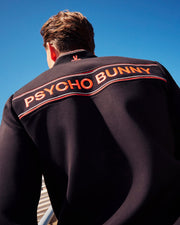 A person with short brown hair is photographed from behind, wearing a MENS COURTSIDE TRACK JACKET - B6J692C200 featuring "PSYCHO BUNNY" in bold orange letters across the upper back. The dark jacket, crafted from stretch cotton-blend jersey, stands out against the clear blue sky.