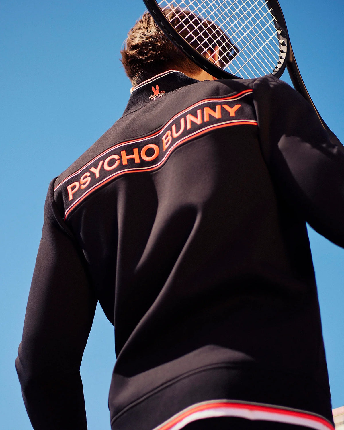 A person wearing a black Psycho Bunny MENS COURTSIDE TRACK JACKET - B6J692C200 holds a tennis racket over their shoulder. The stretch cotton-blend jersey jacket features red and white stripes and the text "PSYCHO BUNNY" on the back. The sky is clear and blue.