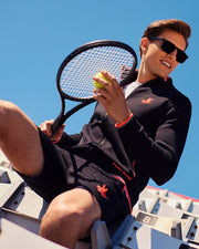 A young man wearing a Psycho Bunny MENS COURTSIDE TRACK JACKET - B6J692C200 with logo-engraved hardware and sunglasses sits on stadium bleachers, holding a tennis racket and ball. His dark athletic outfit with red accents complements the stretch cotton-blend jersey. The sky is clear and blue in the background.