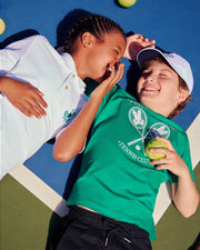 Two children lie on a tennis court, smiling and laughing. One child has their hand partially over their face while whispering to the other. There are tennis balls scattered around them. One wears a green shirt and white cap with the Psycho Bunny logo, while the other sports a white KIDS COURTSIDE GRAPHIC TEE by Psycho Bunny (B0U684C200).