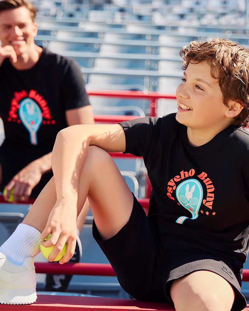 A young boy sits on the bleachers, smiling and holding a tennis ball. He wears the KIDS COURTSIDE GRAPHIC TEE - B0U682C200 by Psycho Bunny, which features a colorful rabbit design. In the background, an adult in a similar tennis-inspired shirt is seated, also holding a tennis ball.