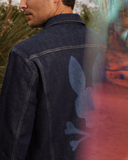 A man wearing the Psycho Bunny MENS PATHE DENIM JACKET - B6J741X1CO, featuring a large stylized bunny and crossbones design on the back, stands outdoors. Blurred colors frame the right side of the image, adding a dynamic effect to this distinctive piece of men's fashion.