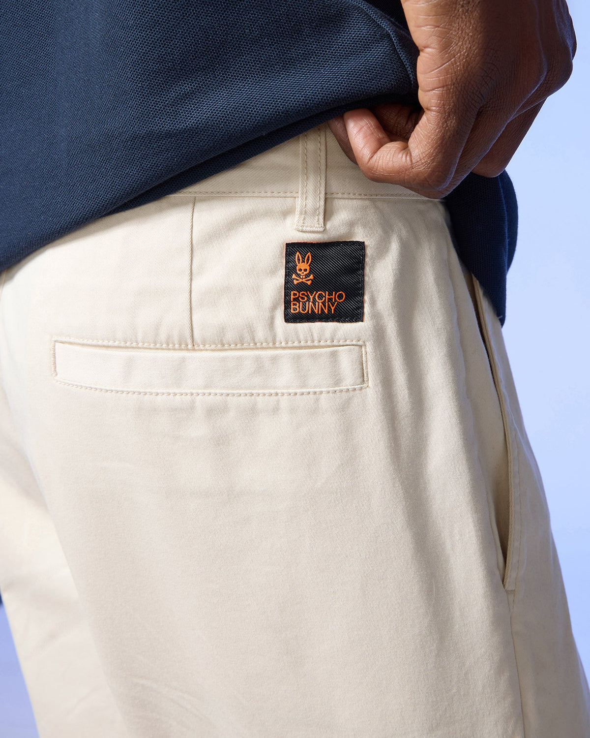A close-up of a person's hand tucked into the back pocket of beige Psycho Bunny Men's York Chino shorts, featuring a black label with a yellow bunny logo and the text "psycho bunny.