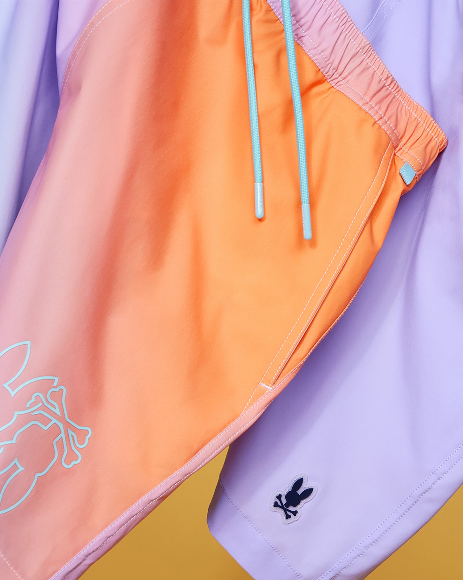 Close-up of two pairs of MENS WINTON PRINTED SWIM TRUNK - B6W915A2PO by Psycho Bunny hanging on a line. One pair is orange with light blue drawstrings and a white rabbit logo. The other pair is light purple with a small black rabbit logo on the leg. Both feature premium finishes and shiny, quick-drying fabric.