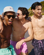 Three men stand on a beach, smiling and laughing. The man on the left wears sunglasses, a white bucket hat, and light blue swim trunks. The man in the middle has curly hair, wears sunglasses, and pink swim trunks. The man on the right sports Psycho Bunny MENS RENDON PRINT SWIM TRUNK - B6W669C200 with a bunny pattern.