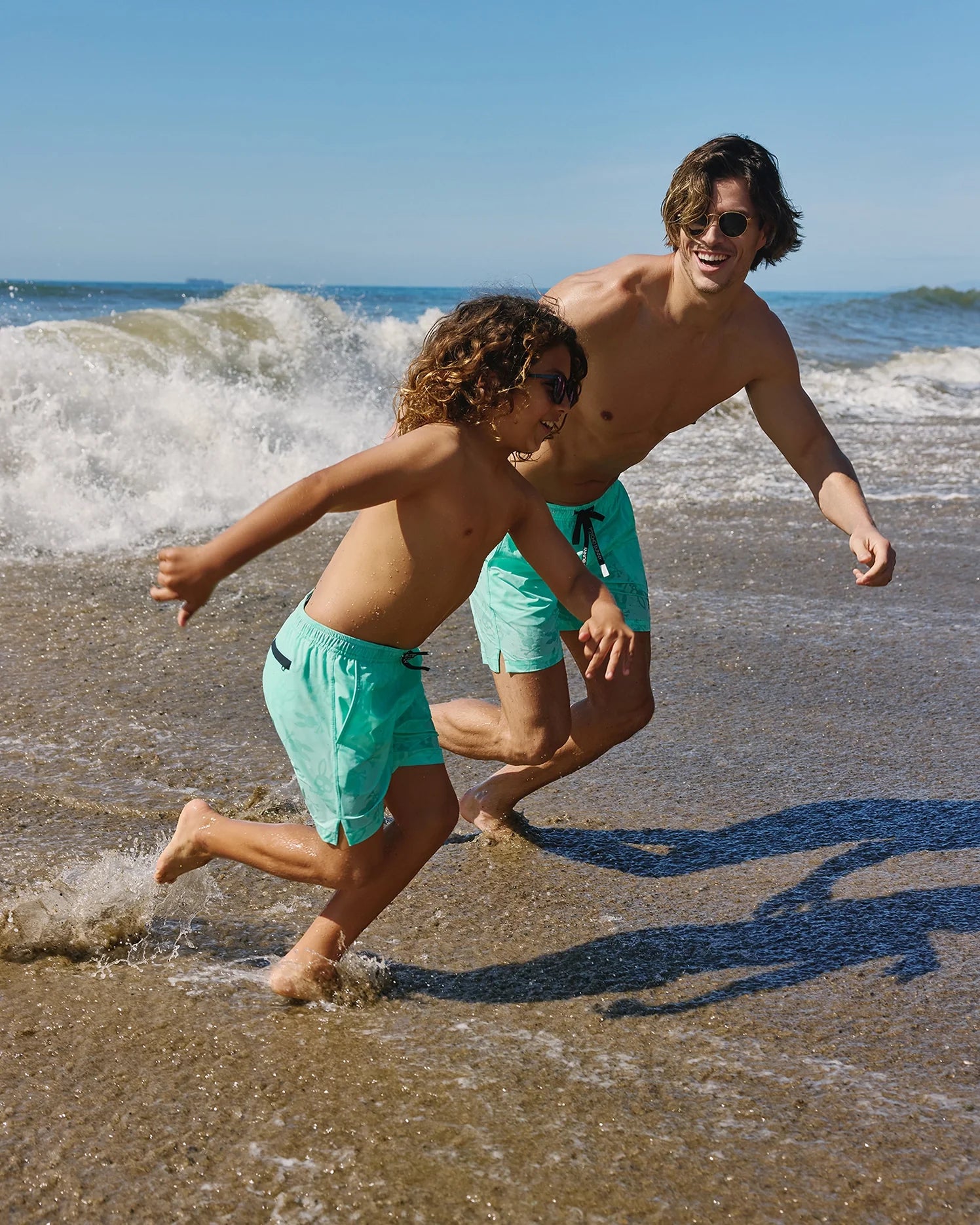 A shirtless adult and child, both sporting Psycho Bunny KIDS PARKER HYDROCHROMIC SWIM TRUNK - B0W646C200 and sunglasses, play and run through shallow waves on a sandy beach. The sky is clear and blue, and they both appear joyful.