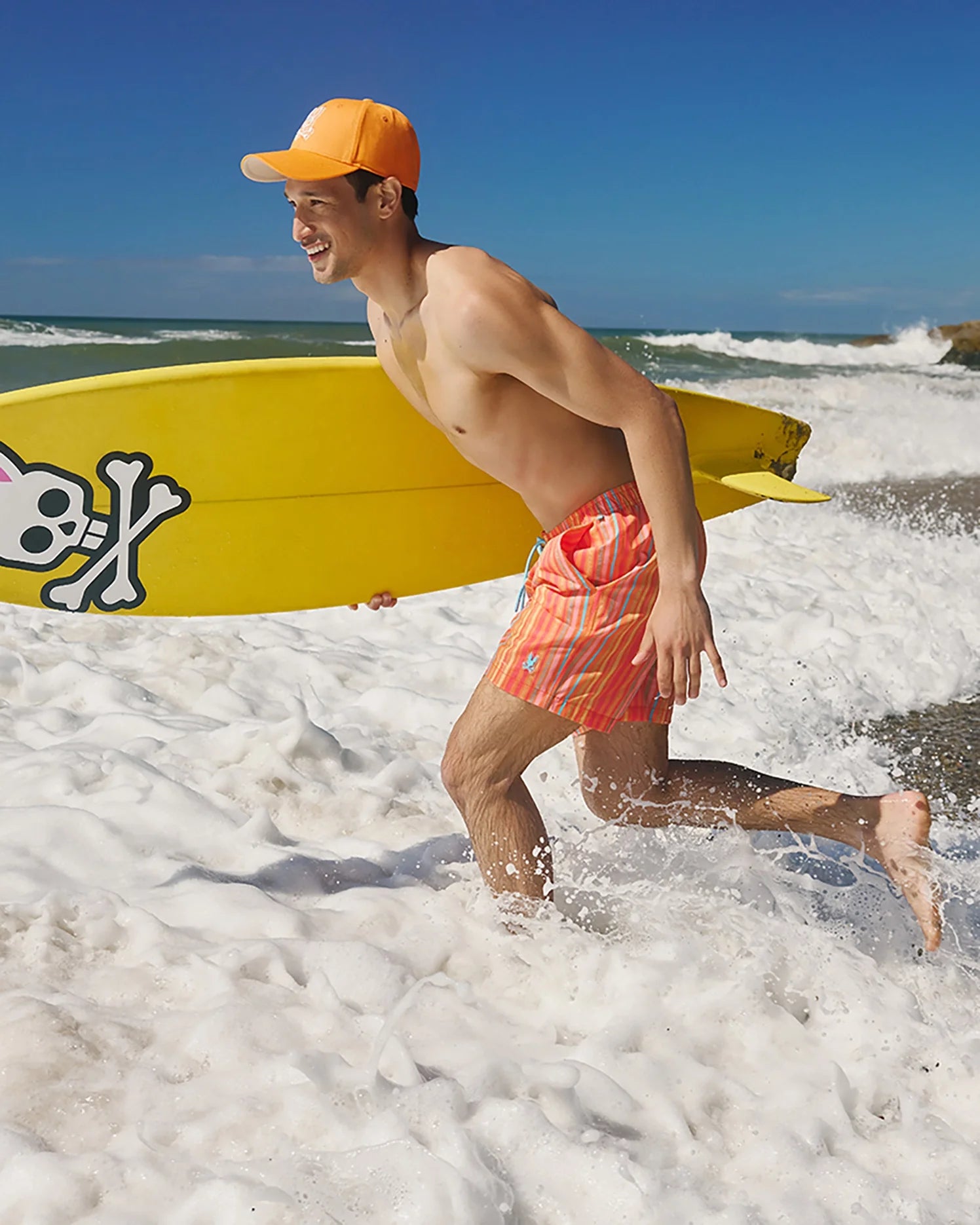 A shirtless man wearing quick-dry orange MENS ANTON ALL OVER PRINT SWIM TRUNK - B6W542C200 from Psycho Bunny and a matching cap holds a yellow surfboard with a skull and crossbones graphic. He is running through foamy waves towards the shore on a sunny beach.