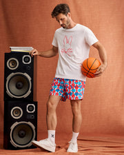 A man stands on a brown backdrop holding an orange basketball in one hand, resting the other on a stack of large black and silver speakers. He is wearing a white T-shirt featuring a pink bunny skull design from Psycho Bunny, MENS WALTER LIGHTWEIGHT PRINT SWIM TRUNK - B6W863D200 with vibrant patterns, white socks, and white sneakers.
