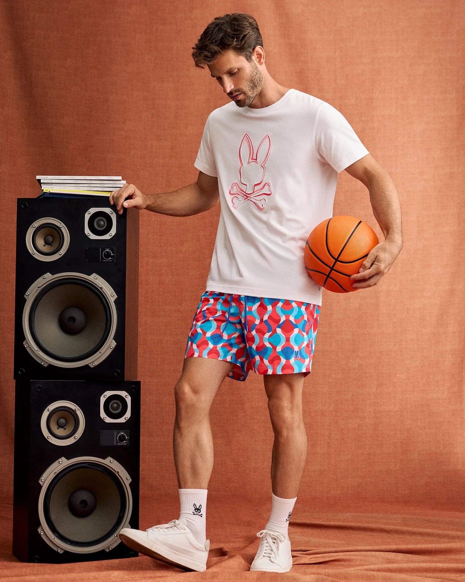 A man stands on a brown backdrop holding an orange basketball in one hand, resting the other on a stack of large black and silver speakers. He is wearing a white T-shirt featuring a pink bunny skull design from Psycho Bunny, MENS WALTER LIGHTWEIGHT PRINT SWIM TRUNK - B6W863D200 with vibrant patterns, white socks, and white sneakers.