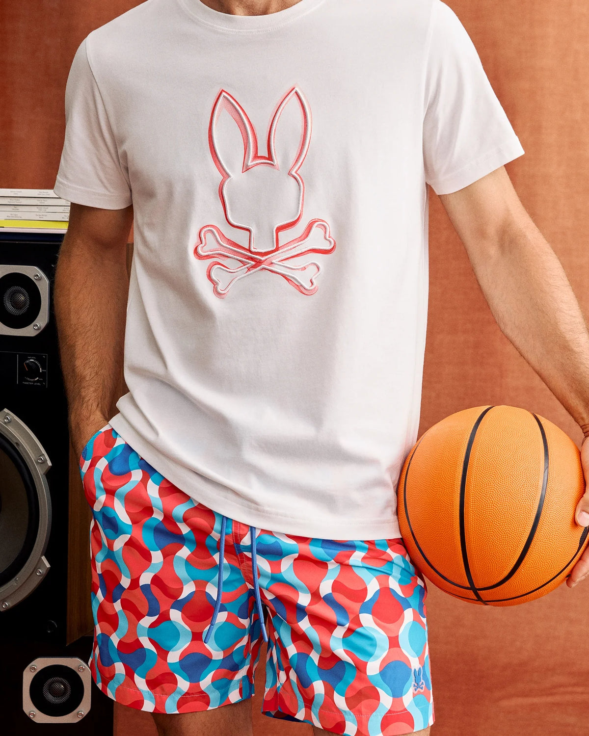 A person wearing a white t-shirt featuring a bunny and crossbones graphic and vibrant Psycho Bunny MENS WALTER LIGHTWEIGHT PRINT SWIM TRUNK (B6W863D200) stands holding an orange basketball. A large black speaker is visible in the background against a brownish backdrop.