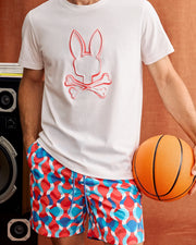 A person wearing a white t-shirt featuring a bunny and crossbones graphic and vibrant Psycho Bunny MENS WALTER LIGHTWEIGHT PRINT SWIM TRUNK (B6W863D200) stands holding an orange basketball. A large black speaker is visible in the background against a brownish backdrop.