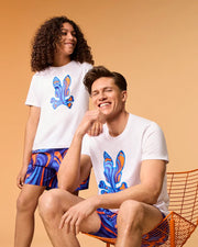 A young person with curly hair and an adult with straight hair are seen sitting and standing against a warm-toned background. Both are dressed in matching outfits, including white T-shirts adorned with colorful abstract rabbit designs, paired with the Psycho Bunny KIDS NEVADA ALL OVER PRINT SWIM TRUNKS (B0W526C200). These multicolored trunks feature quick-dry fabric in blue, orange, and purple patterns.