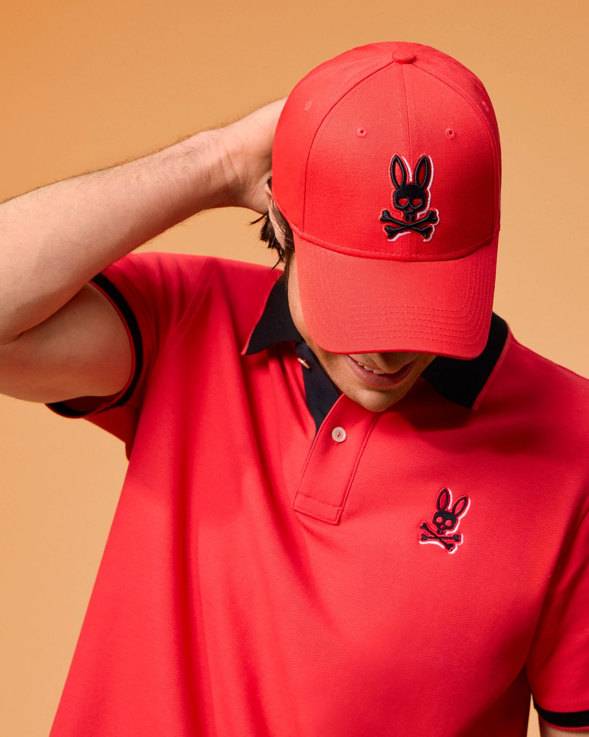 Person wearing a red hat and a Psycho Bunny MENS KAYDEN PIQUE POLO - B6K675C200 with contrasting stripes, featuring a matching bunny skull and crossbones logo. The individual has their hand behind their neck, with their head slightly tilted downwards. The background is a warm beige color.
