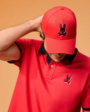 Person wearing a red hat and a Psycho Bunny MENS KAYDEN PIQUE POLO - B6K675C200 with contrasting stripes, featuring a matching bunny skull and crossbones logo. The individual has their hand behind their neck, with their head slightly tilted downwards. The background is a warm beige color.