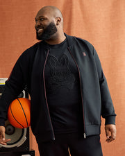 A bearded man smiles while leaning on a speaker with an orange basketball, wearing a BIG AND TALL TRAVIS TRACK JACKET - B9J142D200 by Psycho Bunny over a shirt with a skull and ears design. The backdrop is warm brown.