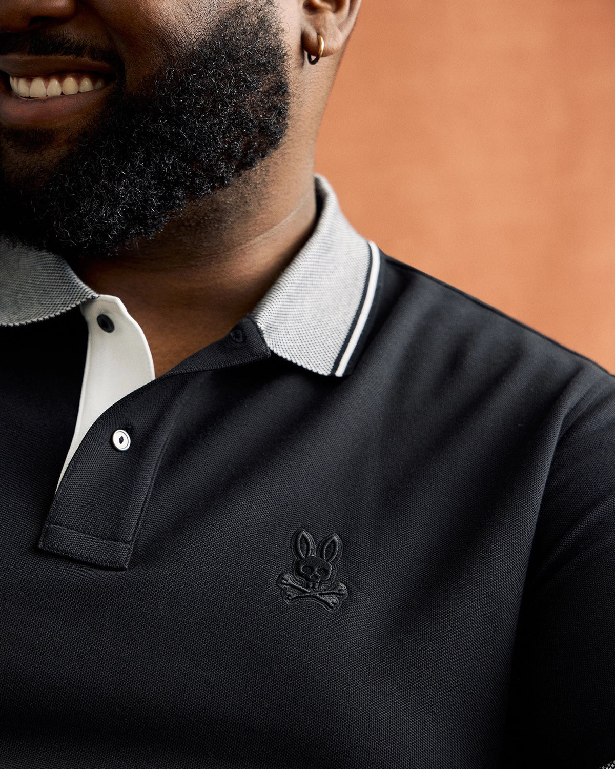 A man wearing a Psycho Bunny BIG AND TALL MALTON PIQUE POLO SHIRT - B9K950D200, with a rabbit logo and white-gray collar trim, stands against a brown background. The soft Pima cotton shirt reveals only the lower half of his bearded face and subtle smile.