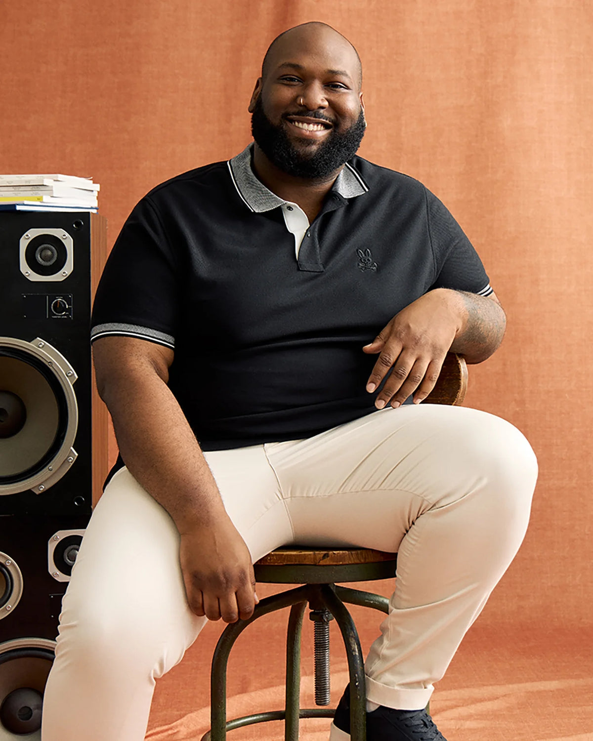 A man smiles while sitting on a stool against a beige backdrop, wearing light pants and a Psycho Bunny Big and Tall Malton Pique Polo Shirt (B9K950D200) in black, with one arm resting on a speaker.