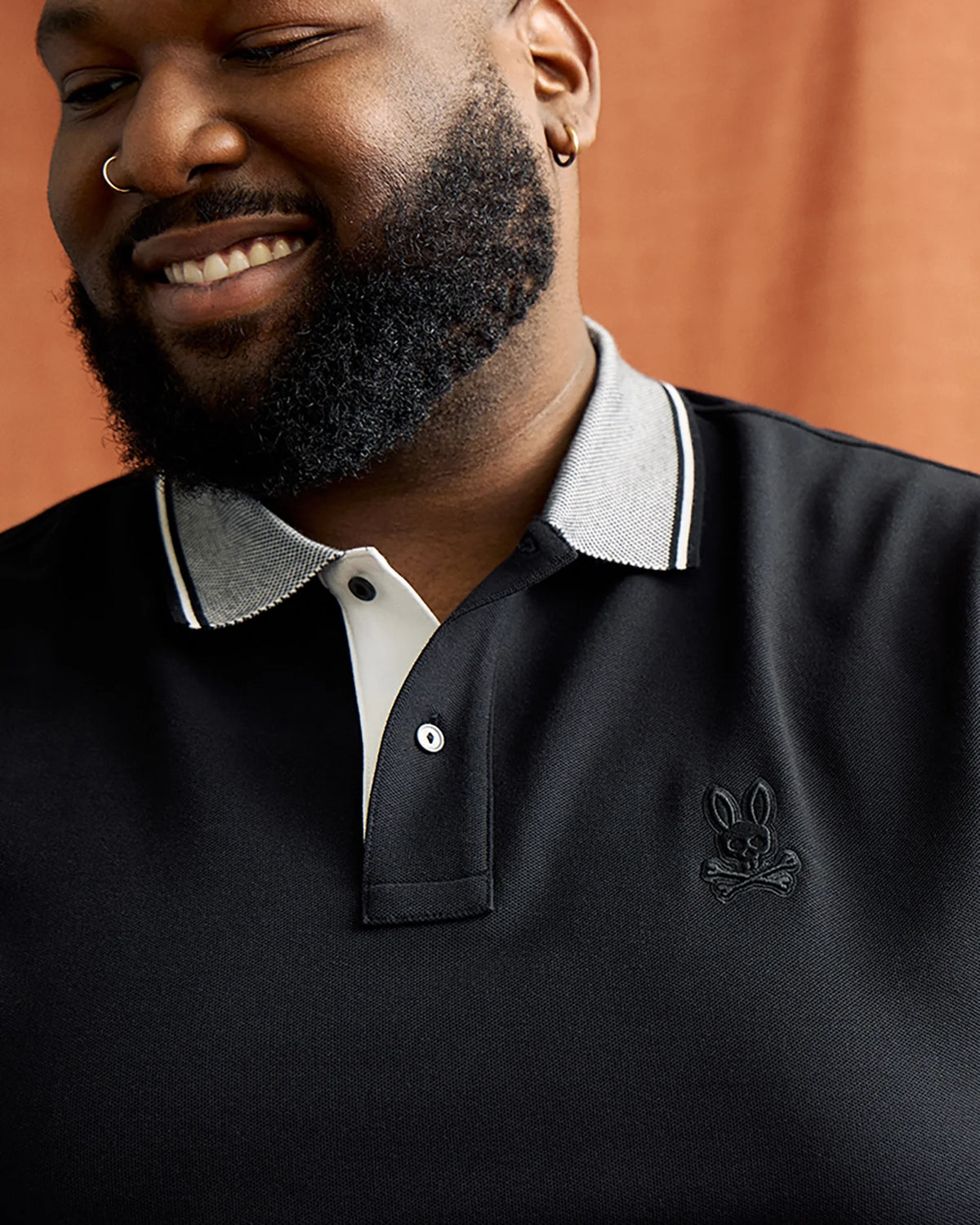 A smiling man with a beard and nose ring wears the black Big and Tall Malton Pique Polo Shirt by Psycho Bunny, featuring a light gray collar and iconic logo, made from luxurious Pima cotton, set against a warm brownish background.