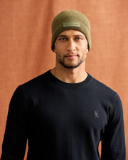 A man wearing a black sweater and the Psycho Bunny MENS SPORT BEANIE - B6A874D200 poses against a warm brown backdrop. With a neutral expression and a slight smile, the beanie is highlighted by its distinctive square patch.