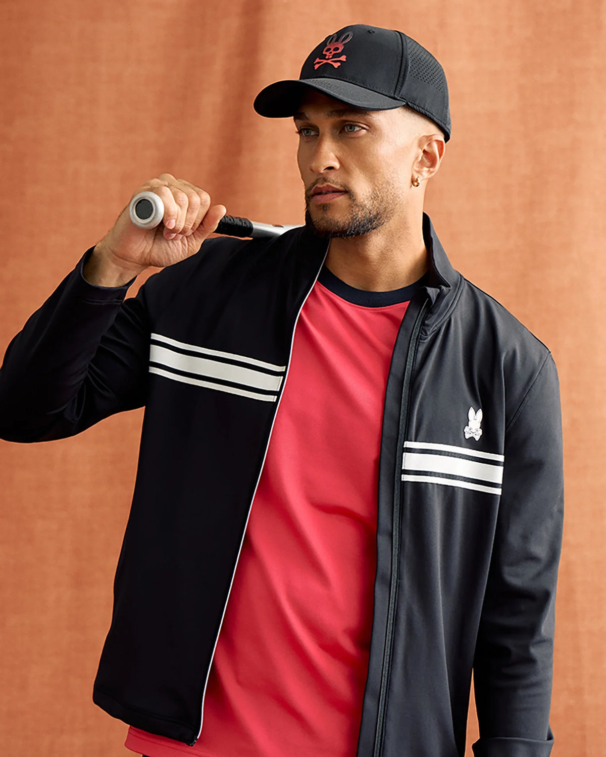 A man wearing the Psycho Bunny MENS TERRELL SPORT ZIP UP JACKET - B6J643C200, which boasts a black and red design with white stripes, holds a tennis racket over his shoulder. He complements the look with a black cap and a red shirt against the warm brown backdrop.