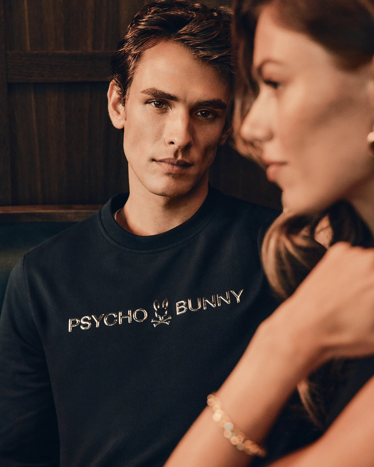 A man in a fashionable black "Mens Lavata French Terry Sweatshirt" from Psycho Bunny gazes forward, seated next to a woman with blurred facial features. The background is out of focus, creating an intimate, moody atmosphere.