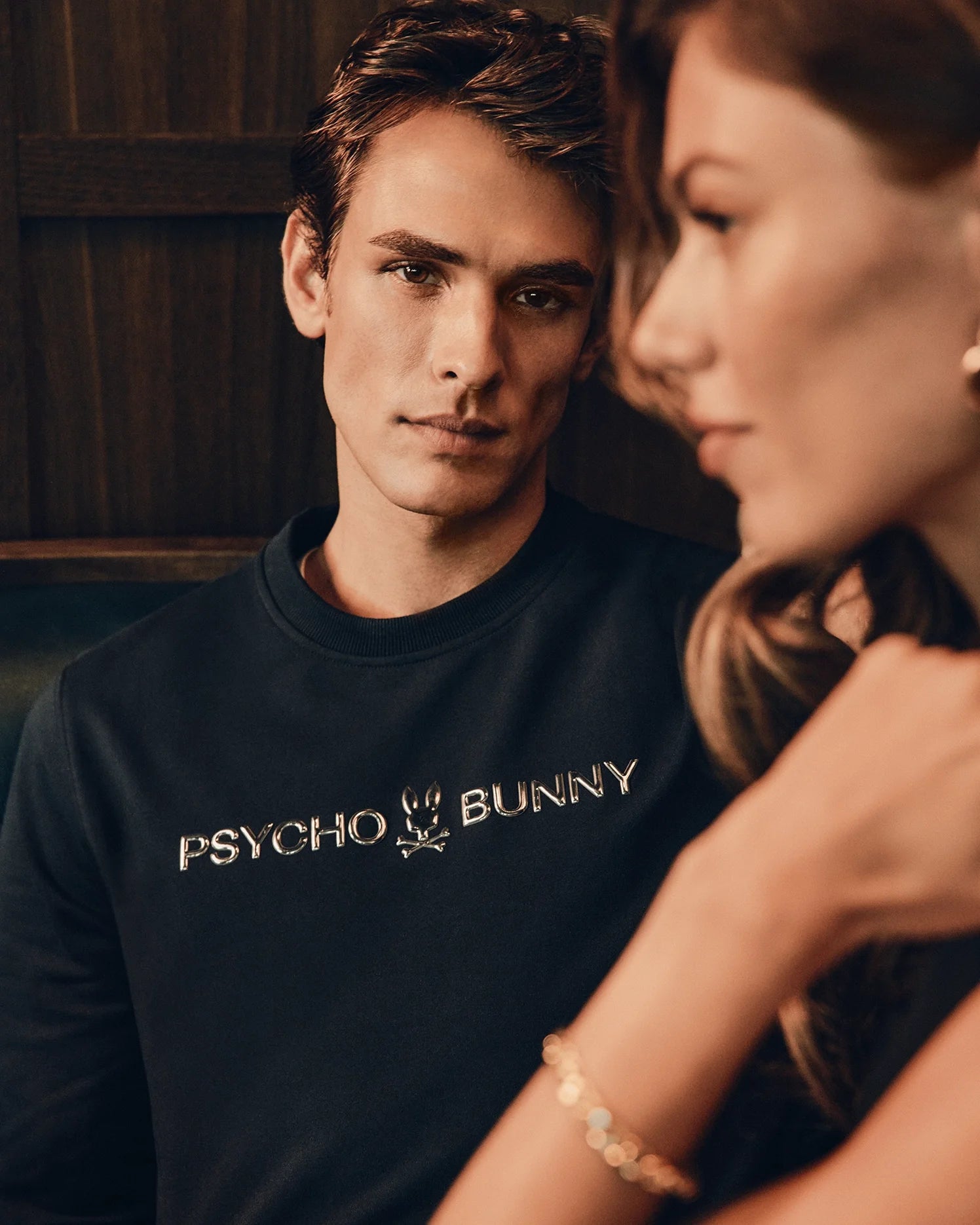 A man in a fashionable black "Mens Lavata French Terry Sweatshirt" from Psycho Bunny gazes forward, seated next to a woman with blurred facial features. The background is out of focus, creating an intimate, moody atmosphere.