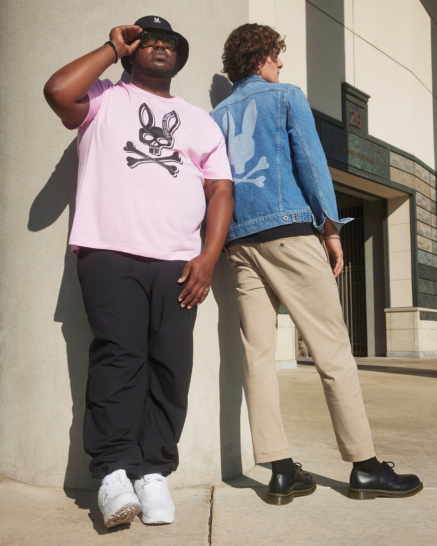 Two people stand by a building. One wears a pink T-shirt, black pants, white sneakers, and a black hat. The other sports a denim jacket with rabbit imagery and Psycho Bunny's MENS DELANCY CHINO PANTS in beige with black shoes. Both feature rabbit designs on their outfits.