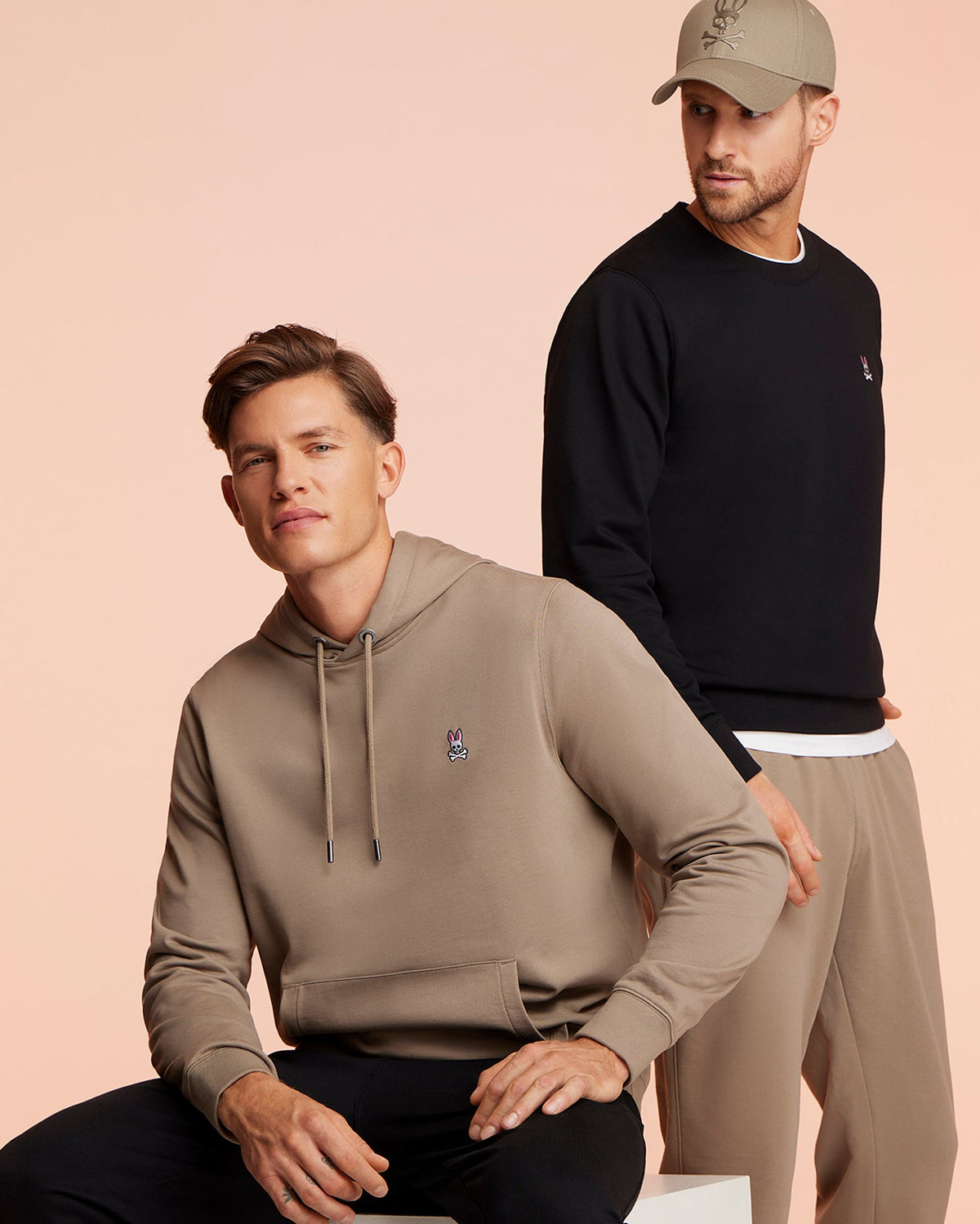 Two men model against a peach backdrop: one in a beige hoodie with classic black pants, the other in a black sweatshirt, Psycho Bunny beige French terry sweatpants (B6P828ARFT), and matching cap. Both outfits boast a small embroidered logo.