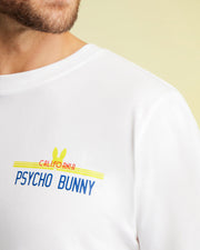 A close-up displays an individual donning a breathable white MENS CALIFORNIA TEE - B6U552W1PC by Psycho Bunny, with "California Psycho Bunny" emblazoned in blue and red. The design includes a yellow bunny silhouette behind the text. The focus is on the graphic, highlighting the smooth texture of the Pima cotton jersey while partially revealing the person's face.