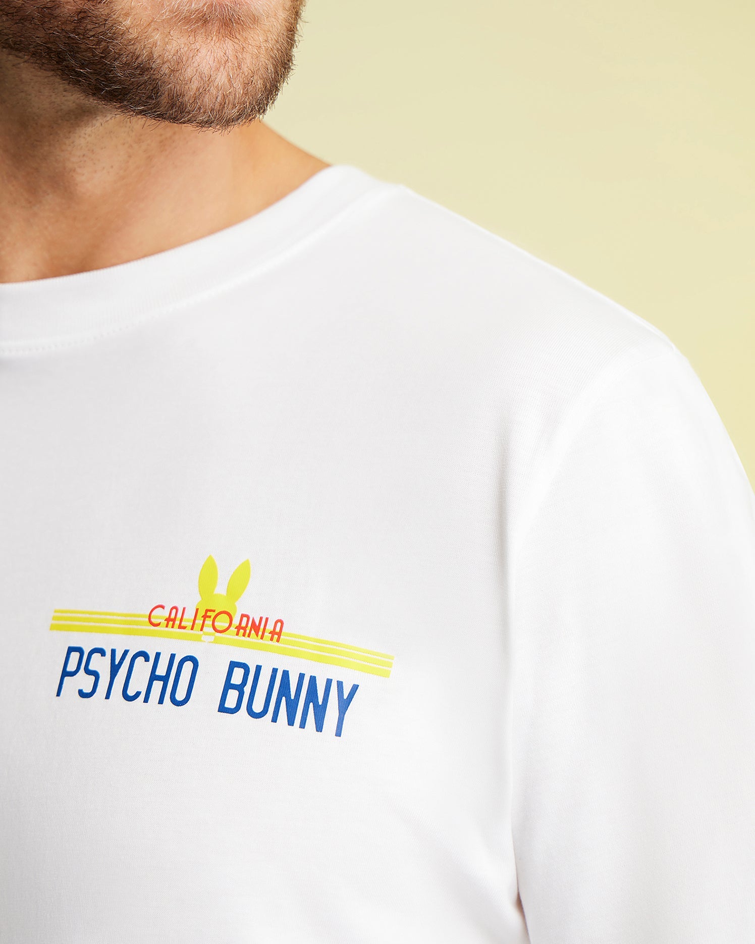 A close-up displays an individual donning a breathable white MENS CALIFORNIA TEE - B6U552W1PC by Psycho Bunny, with "California Psycho Bunny" emblazoned in blue and red. The design includes a yellow bunny silhouette behind the text. The focus is on the graphic, highlighting the smooth texture of the Pima cotton jersey while partially revealing the person's face.