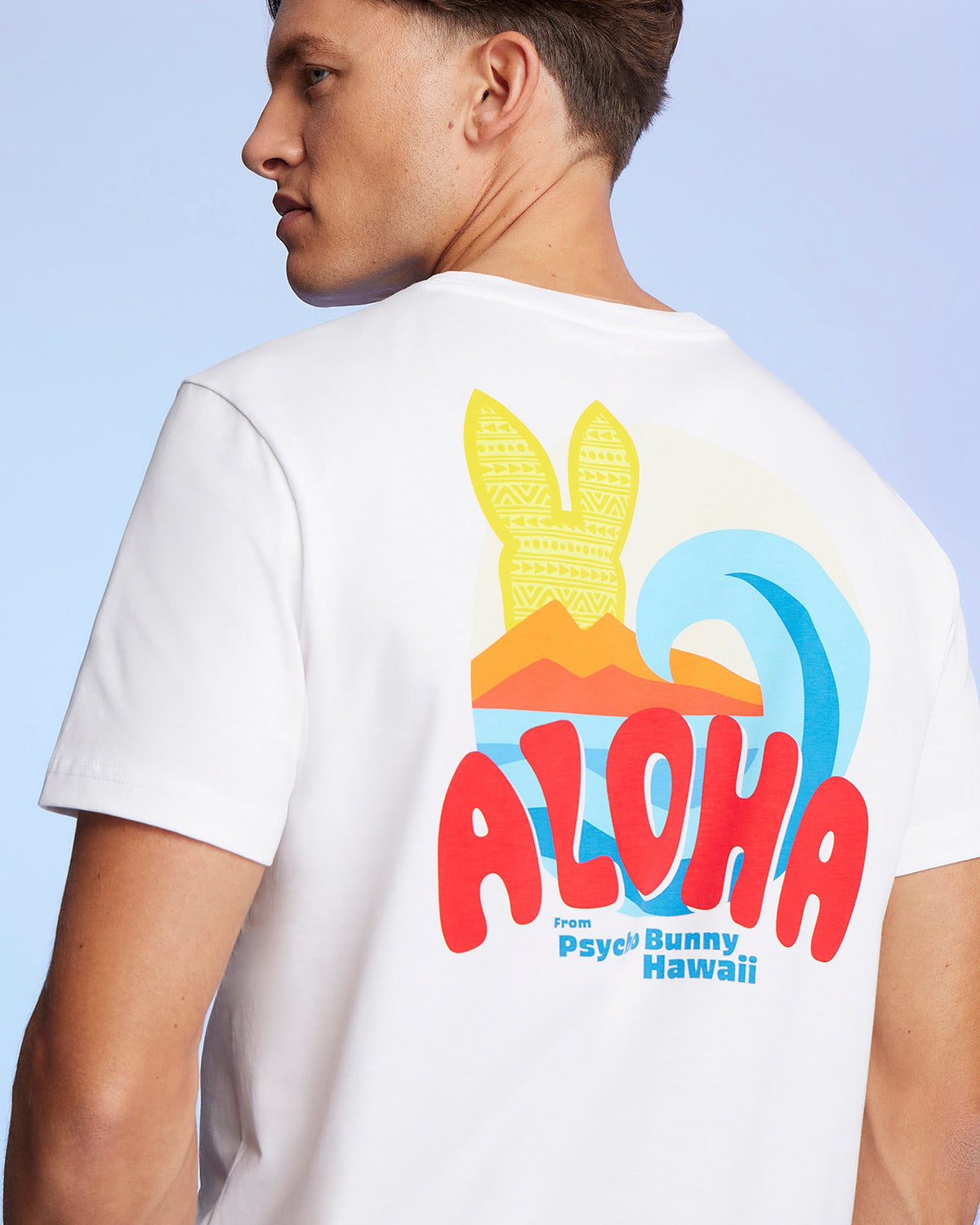 A man is shown from the side wearing a white Pima cotton jersey MENS HAWAII CITY TEE - B6U169Y1PC with a colorful design on the back. The design features the text "ALOHA" in red letters, a beach scene with a wave, sun, and surfboards, and additional text "from Psycho Bunny Hawaii.
