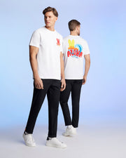 Two men are standing against a light blue background, wearing matching white t-shirts and black pants. The man on the left faces the camera, showcasing a red graphic pattern on his Pima cotton jersey. The man on the right is turned away, revealing a colorful "ALOHA" Hawaii tee design on the back of his MENS HAWAII CITY TEE - B6U169Y1PC by Psycho Bunny.