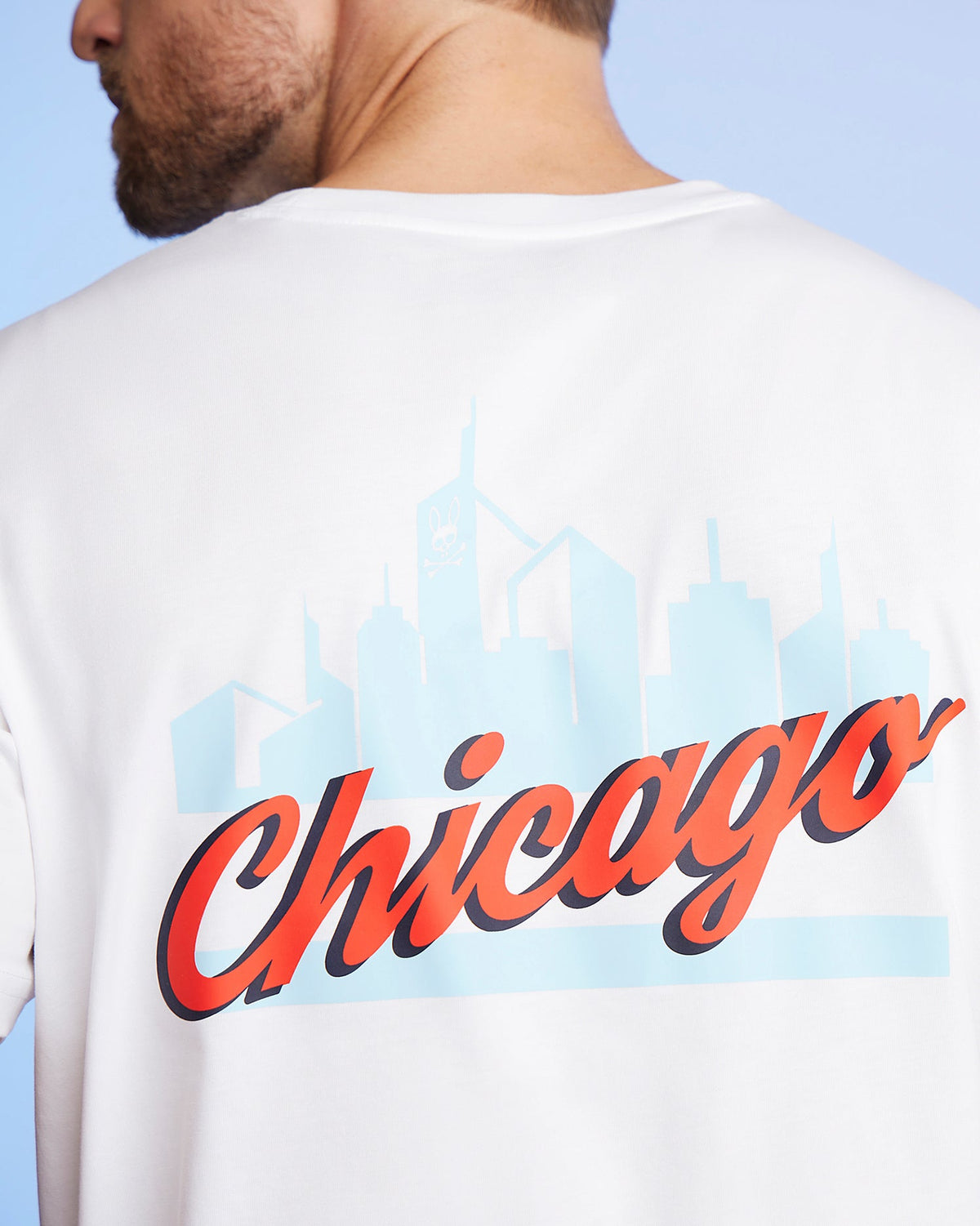 The MENS CHICAGO CITY TEE - B6U551W1PC by Psycho Bunny showcases a close-up of someone wearing this stylish tee, made from soft Pima cotton jersey. It features "Chicago" boldly written in red script across the back, with a light blue city skyline silhouette in the background and a subtle bunny logo graphic.