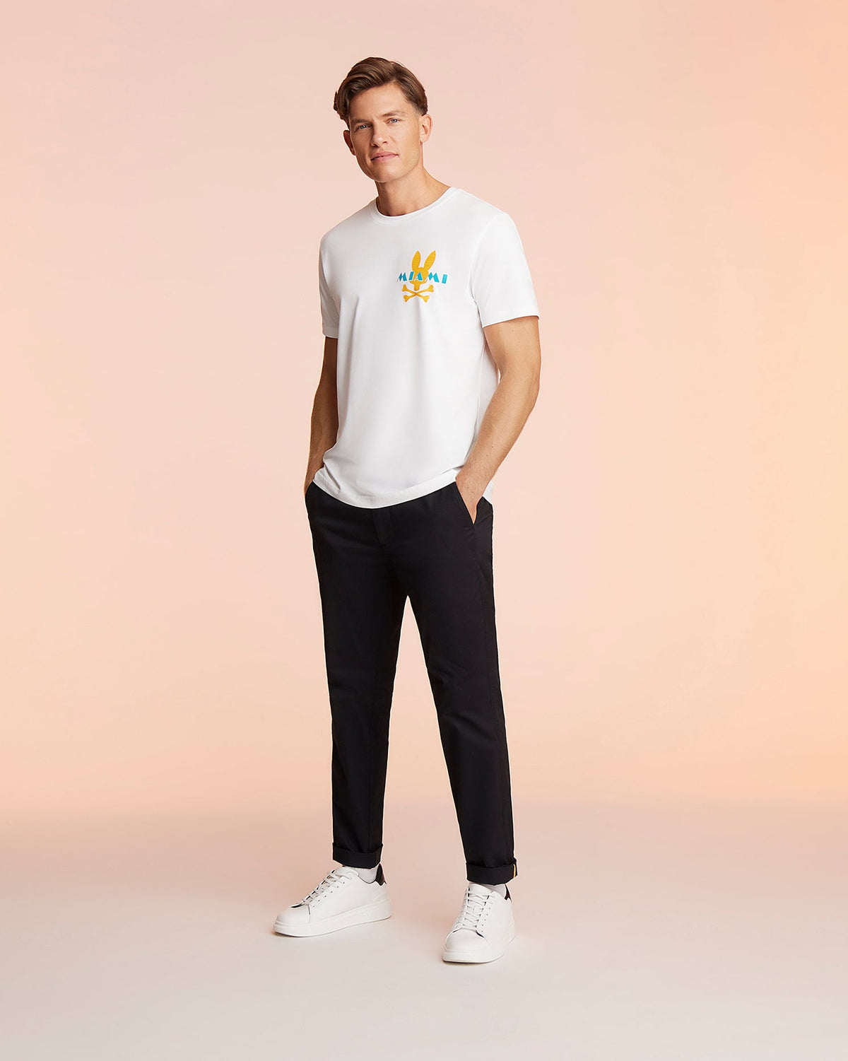 A man stands against a light peach background, sporting a white Pima cotton Psycho Bunny MENS MIAMI TEE - B6U550W1PC with a colorful Miami graphic on it, black pants, and white sneakers. His hands are in his pockets as he looks slightly off to the side.