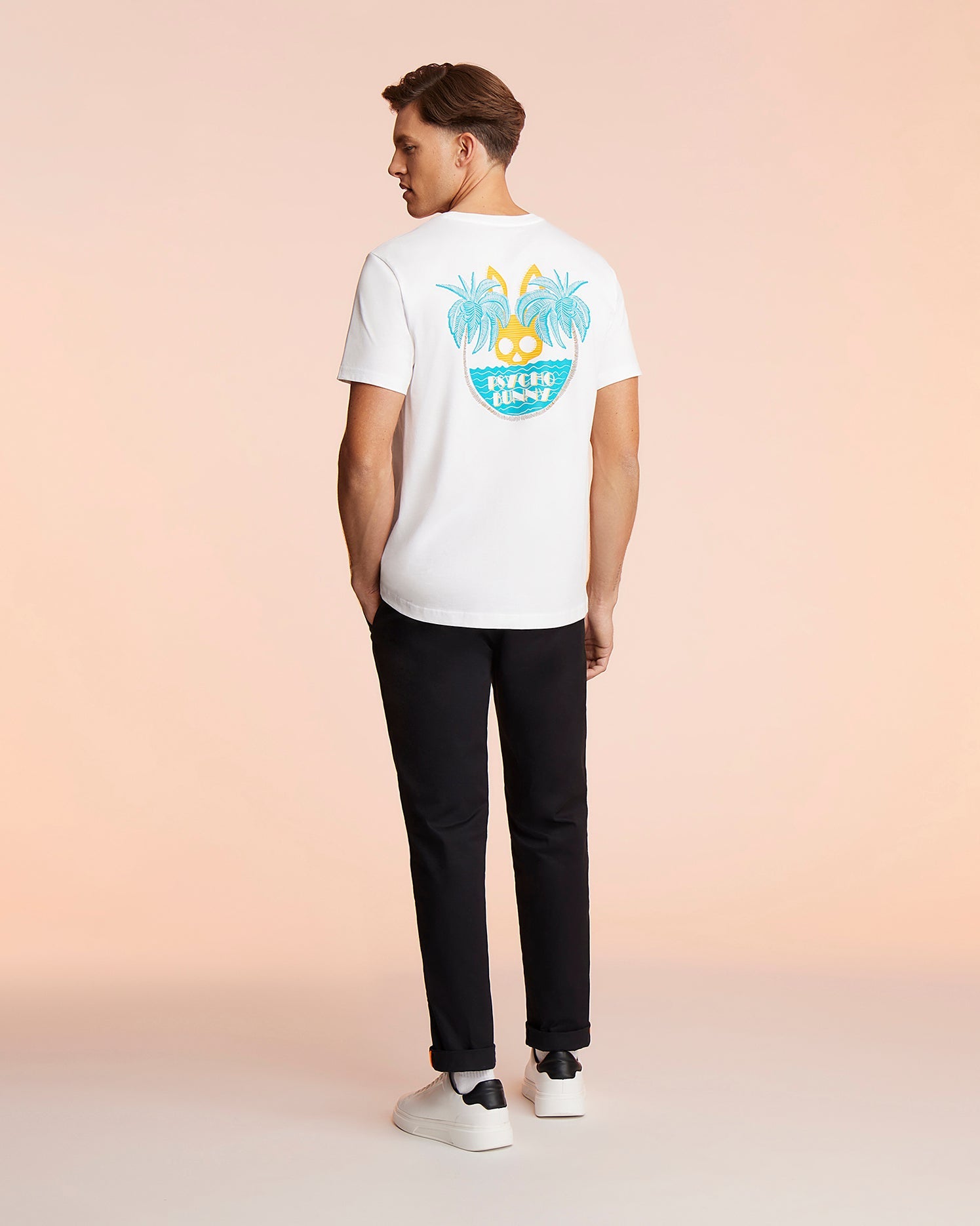 A man stands facing away, wearing a white Psycho Bunny MENS MIAMI TEE - B6U550W1PC made of soft Pima cotton with a colorful tropical design featuring palm trees and a sunset on the back, paired with black pants and white sneakers. The background is a soft peach color, evoking the vibrant vibe of Miami.