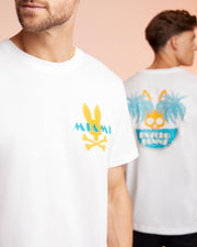 Two men wearing white men's Florida tees with colorful graphics. The man in the foreground has a Psycho Bunny MENS MIAMI TEE - B6U550W1PC that says "Miami" with a yellow bunny logo. The man in the background has a graphic of palm trees and text on his T-shirt’s back.