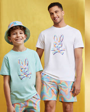 A boy and a man stand smiling in front of a yellow background. Both are wearing matching clothing featuring a colorful, holographic bunny and crossbones design. The boy wears a mint green t-shirt, quick-drying swim trunks, and a matching bucket hat. The man wears a white t-shirt and similar shorts from Psycho Bunny's MENS BLOOMINGTON SWIM TRUNK - B6W941A2PO collection.