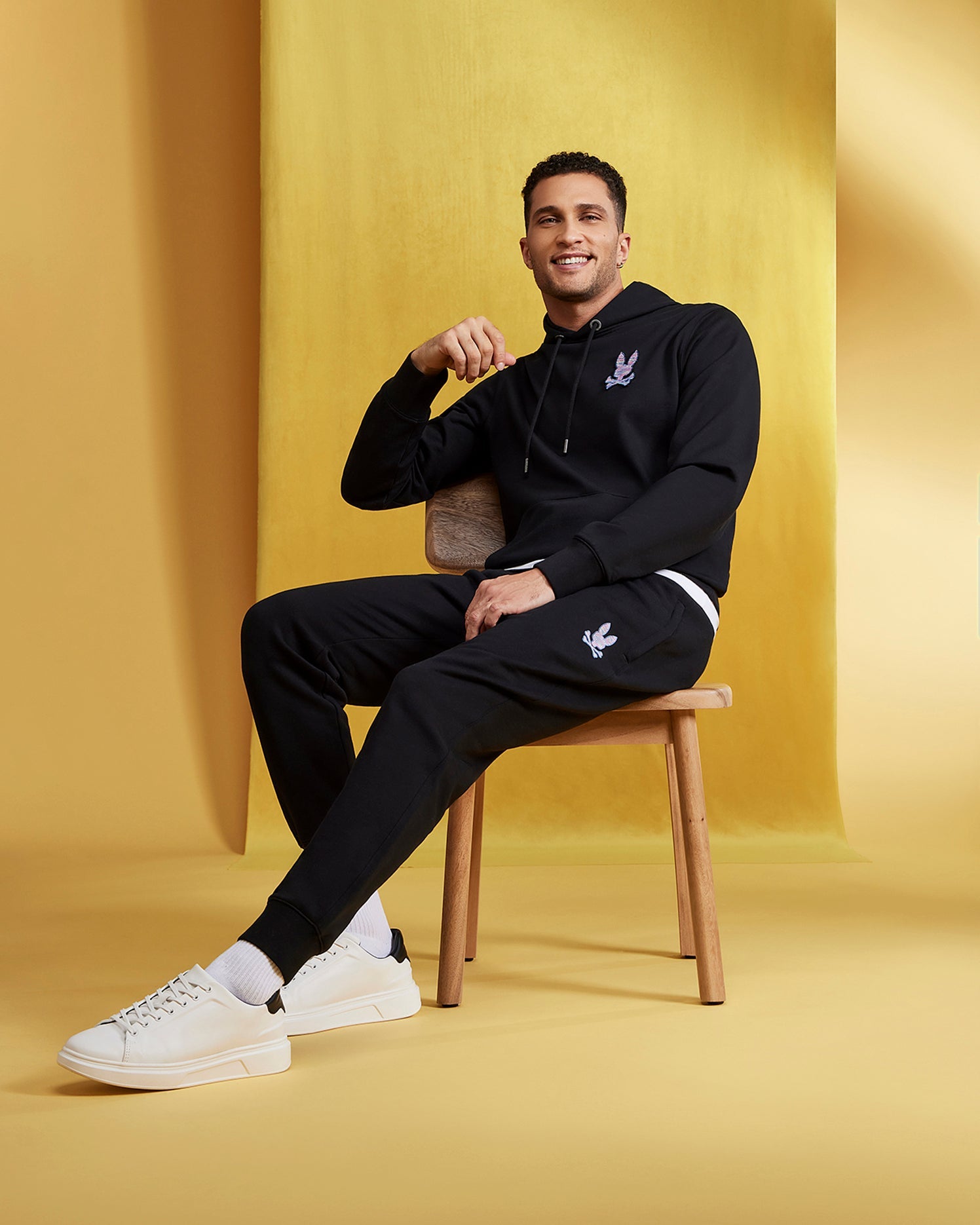 A smiling man is seated on a wooden chair against a yellow backdrop. He is wearing a black hoodie and Psycho Bunny MENS SANTA CRUZ SWEATPANT - B6P843A2FT with a white emblem, paired with white sneakers. Crafted from 100% cotton, these sweatpants offer both comfort and style. His right hand rests near his chin, and his left hand is on his knee.