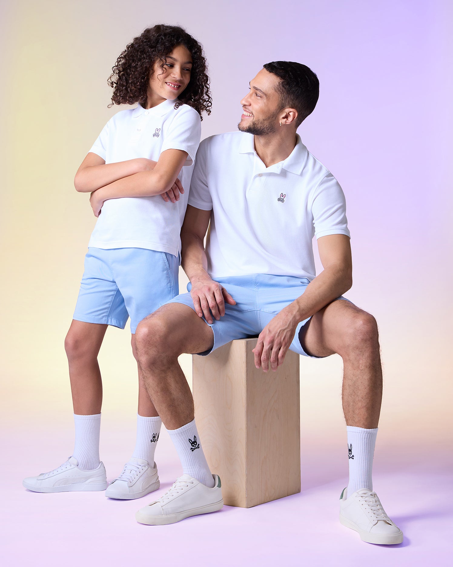 A woman and a man wearing casual sporty attire, including white polo shirts with embroidered Psycho Bunny logos and light blue 
MENS WILLIS STRETCH TENCEL SHORT - B6R239Y1WB, posing with smiles in a studio with a pastel-colored background.