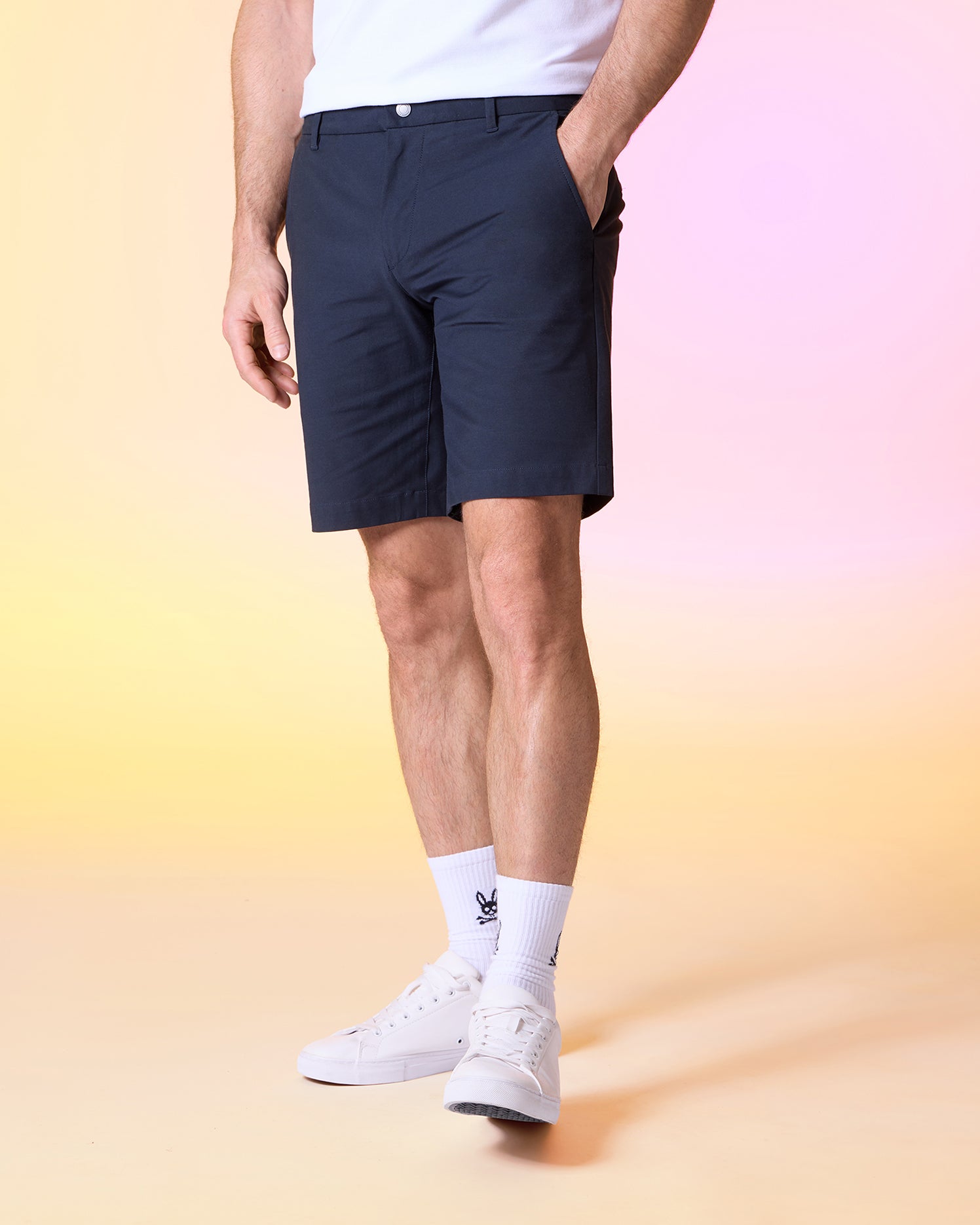 A person wearing dark blue Psycho Bunny MENS GABLE REGULAR FIT SPORT SHORT - B6R761ARCN made from durable stretch cotton-nylon and a white shirt, with their hands in their pockets. They also have white socks and white sneakers on. The background is a gradient, transitioning from yellow at the bottom to pink at the top.