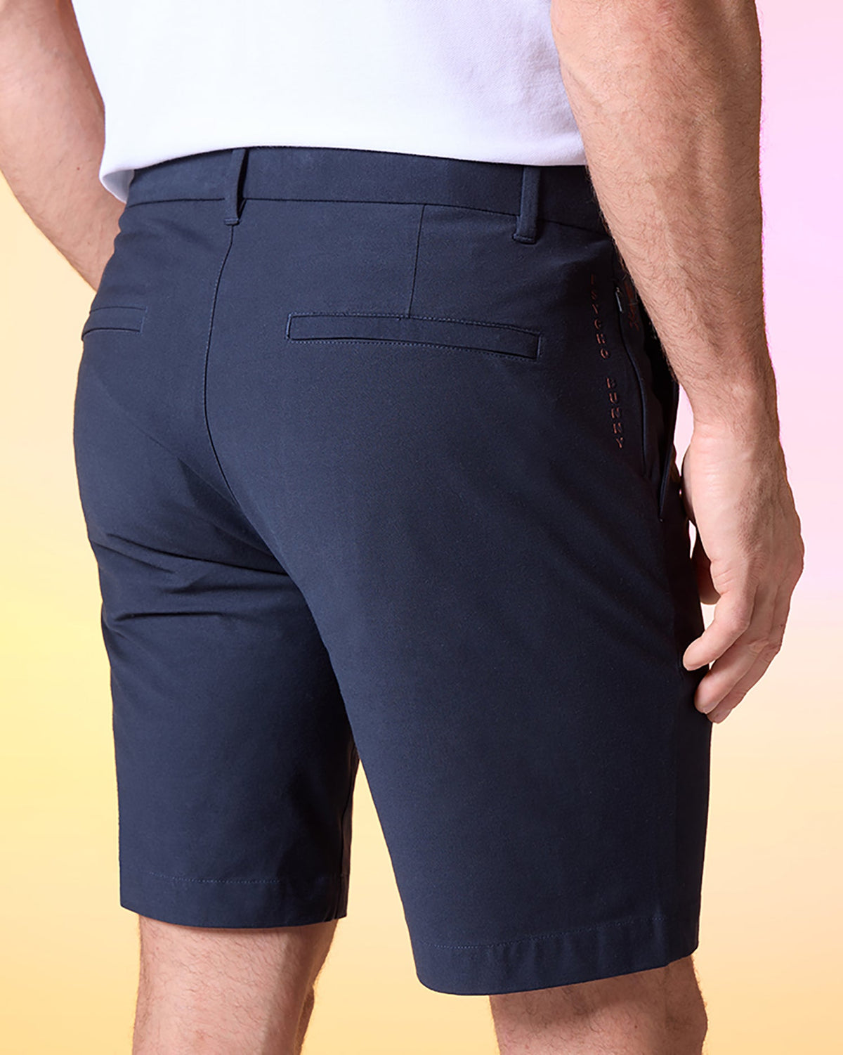 A man wearing a white shirt and navy blue Psycho Bunny MENS GABLE REGULAR FIT SPORT SHORT - B6R761ARCN stands with his back facing the camera. The durable stretch cotton-nylon shorts have belt loops, two back pockets with horizontal slits, a hidden zip pocket, and a small stitched design on the right side seam. The background is a gradient of yellow and pink.
