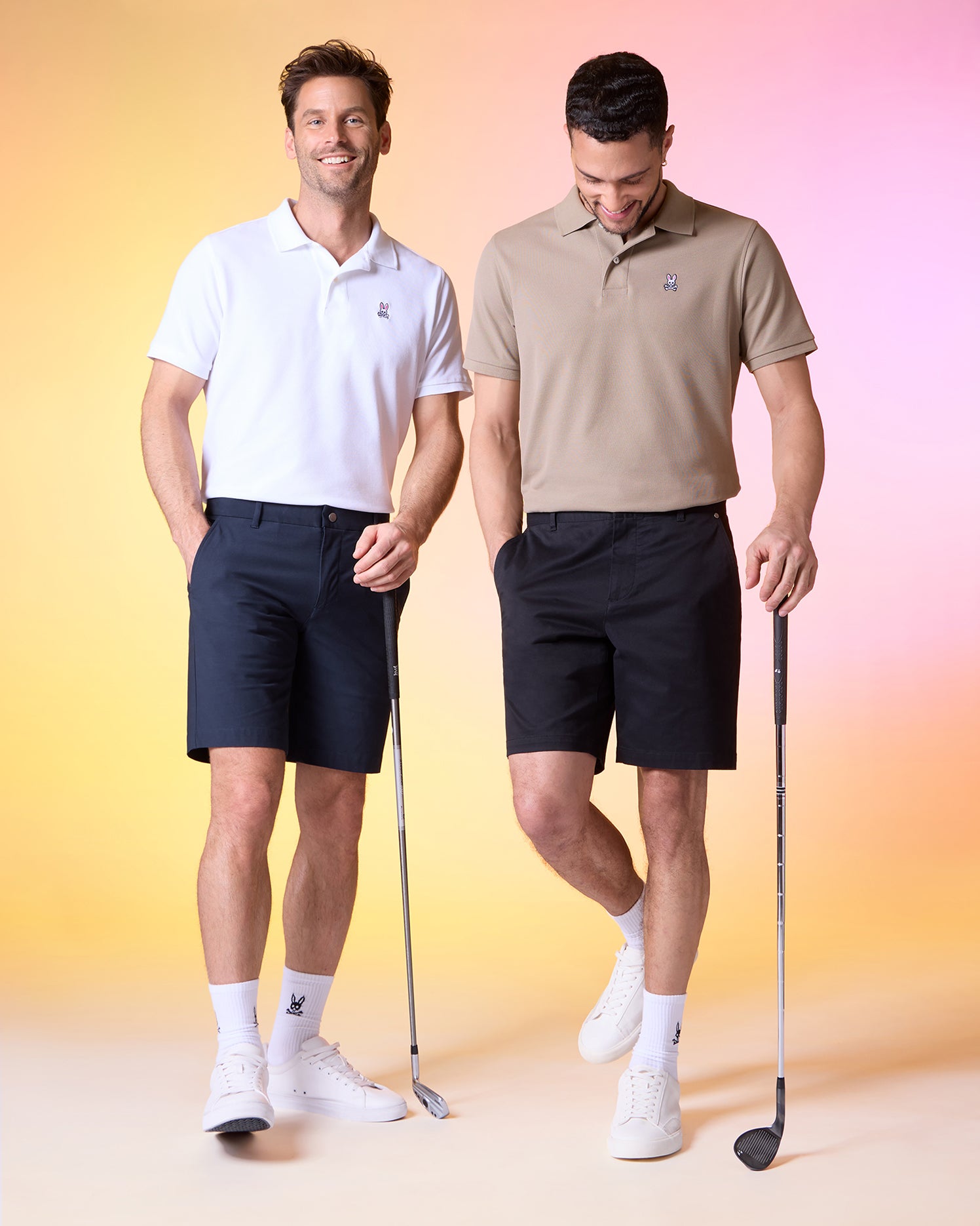 Two men in casual golf attire pose against a gradient background. Both wear polo shirts, shorts, and sneakers; the man on the left wears a white shirt and black MENS GABLE REGULAR FIT SPORT SHORT - B6R761ARCN by Psycho Bunny with a hidden zip pocket, while the man on the right sports durable stretch cotton-nylon tan shirt and black shorts, holding a golf club.