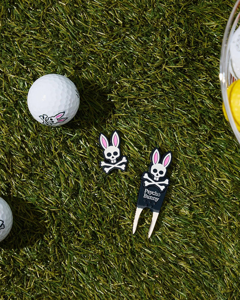 Image shows golf gifts on grass: a golf ball with a bunny skull logo, a black DIVOT TOOL - B6A699C200 featuring the text "Psycho Bunny" and two magnetic ball markers with the same logo. A bowl of yellow and white golf balls is partly visible.