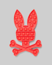 The PSYCHO BUNNY POP IT - B0A376E200 by Psycho Bunny features a red bunny head with long ears above crossed bones, resembling a fun skull-and-crossbones. Made from high-quality silicone for stress relief, set against a light gray background.