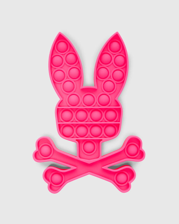 The PSYCHO BUNNY POP IT - B0A376E200 by Psycho Bunny is a bunny-shaped pop-it fidget toy made from premium silicone, featuring a pink rabbit head with long ears atop crossed bones in a skull-and-crossbones design.