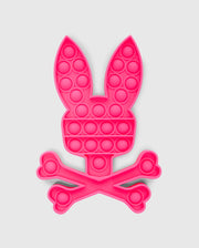 The PSYCHO BUNNY POP IT - B0A376E200 by Psycho Bunny is a bunny-shaped pop-it fidget toy made from premium silicone, featuring a pink rabbit head with long ears atop crossed bones in a skull-and-crossbones design.