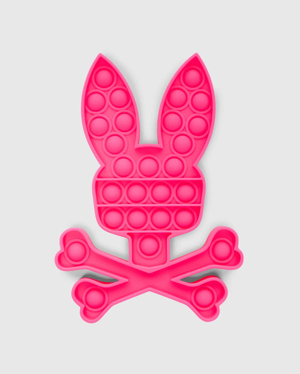 The PSYCHO BUNNY POP IT - B0A376E200 by Psycho Bunny is a bunny-shaped pop-it fidget toy made from premium silicone, featuring a pink rabbit head with long ears atop crossed bones in a skull-and-crossbones design.